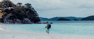 Person walking on a sandy beach with clear blue water and mountains in the background